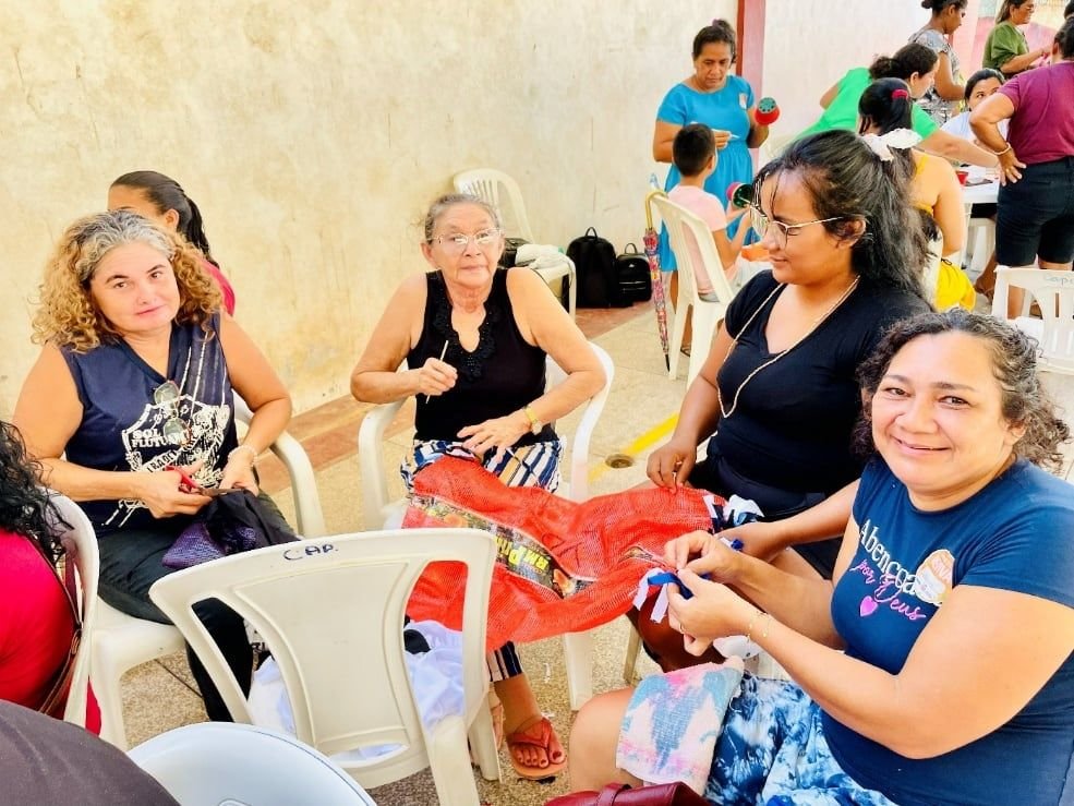 Mulheres em roda trabalhando com sacos de rede vermelha, uma delas sorrindo para a câmera