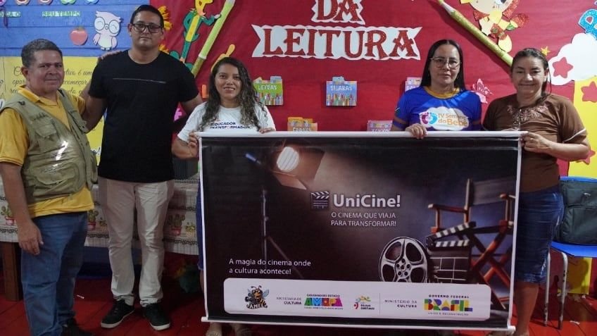 Pessoas segurando o banner do UniCine diante de um painel do Ateliê da Leitura