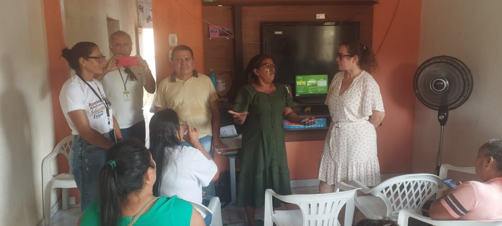 Pessoas reunidas numa sala, uma delas falando ao lado de uma televisão